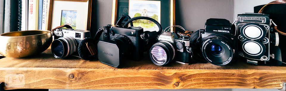 A collection of vintage and modern cameras displayed on a wooden shelf, alongside a brass bowl.