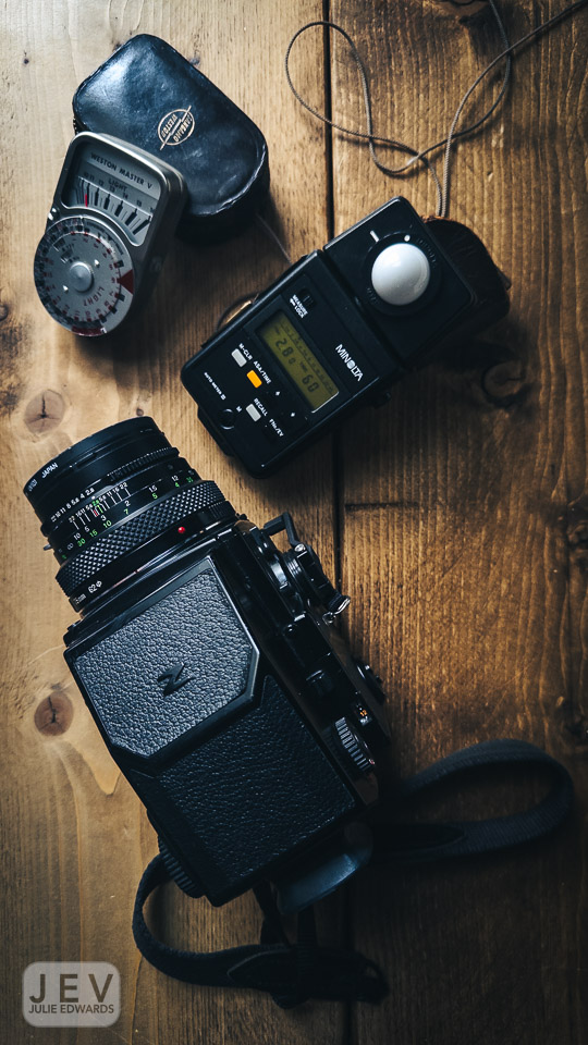 An overhead view of a camera with a black textured body and a lens, accompanied by a light meter and a vintage exposure meter on a wooden surface.